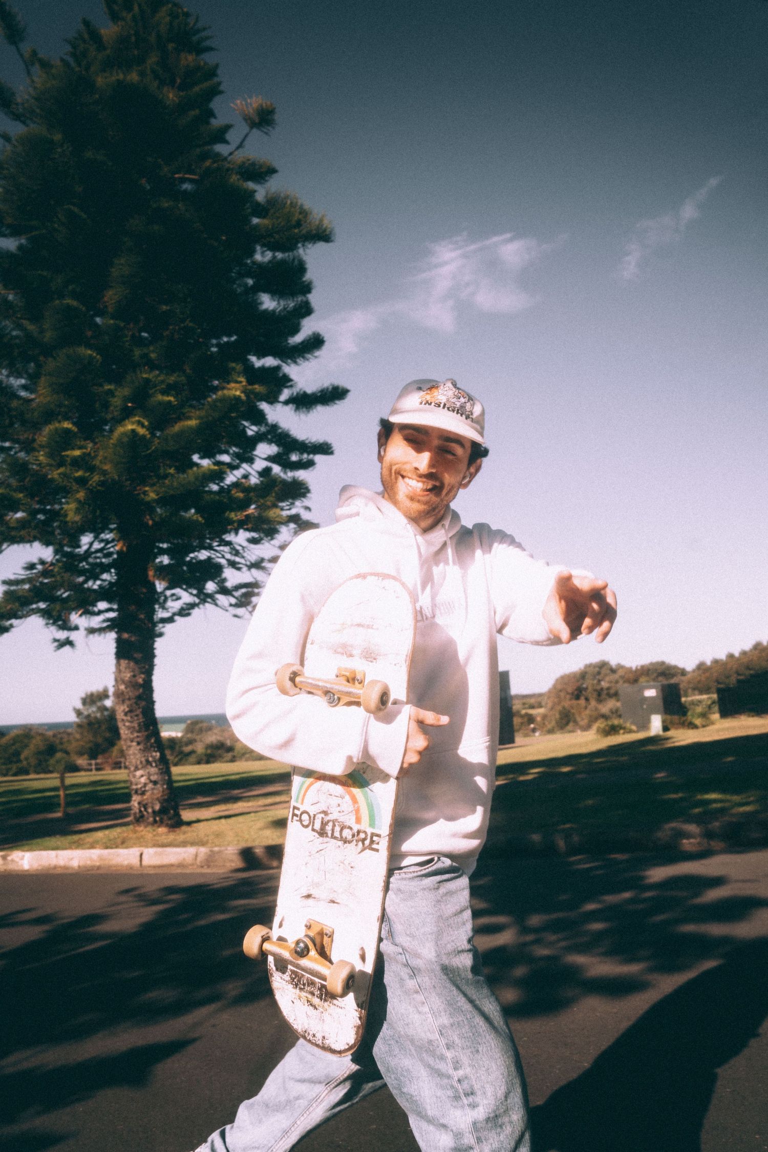 Casual skater holding a skateboard while standing near a pine tree on a sunny day.