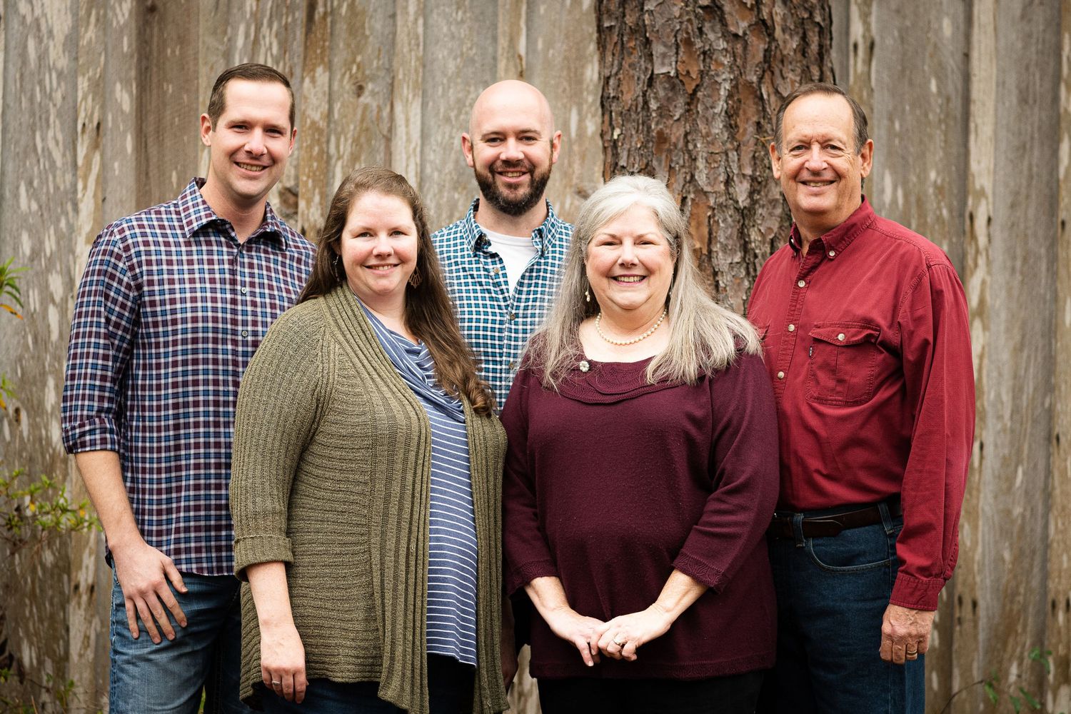 Multi-generational family photo taken against weathered wood barn wall with tall pine tree.