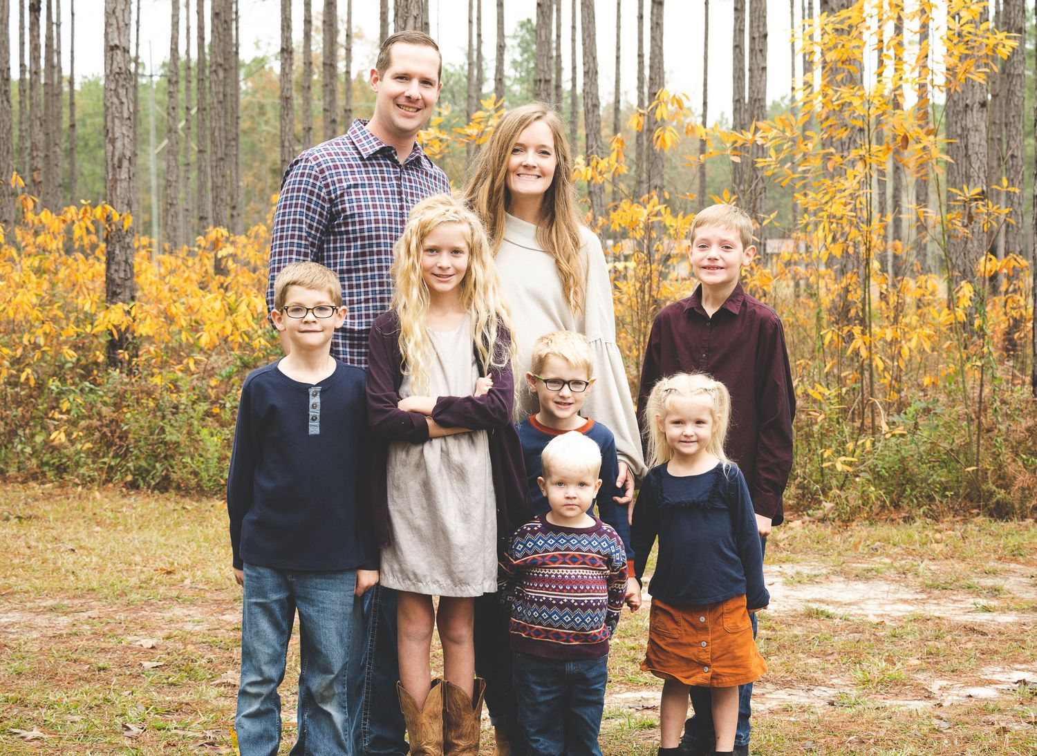 Family group photo outdoors with pine trees and golden fall foliage in background.