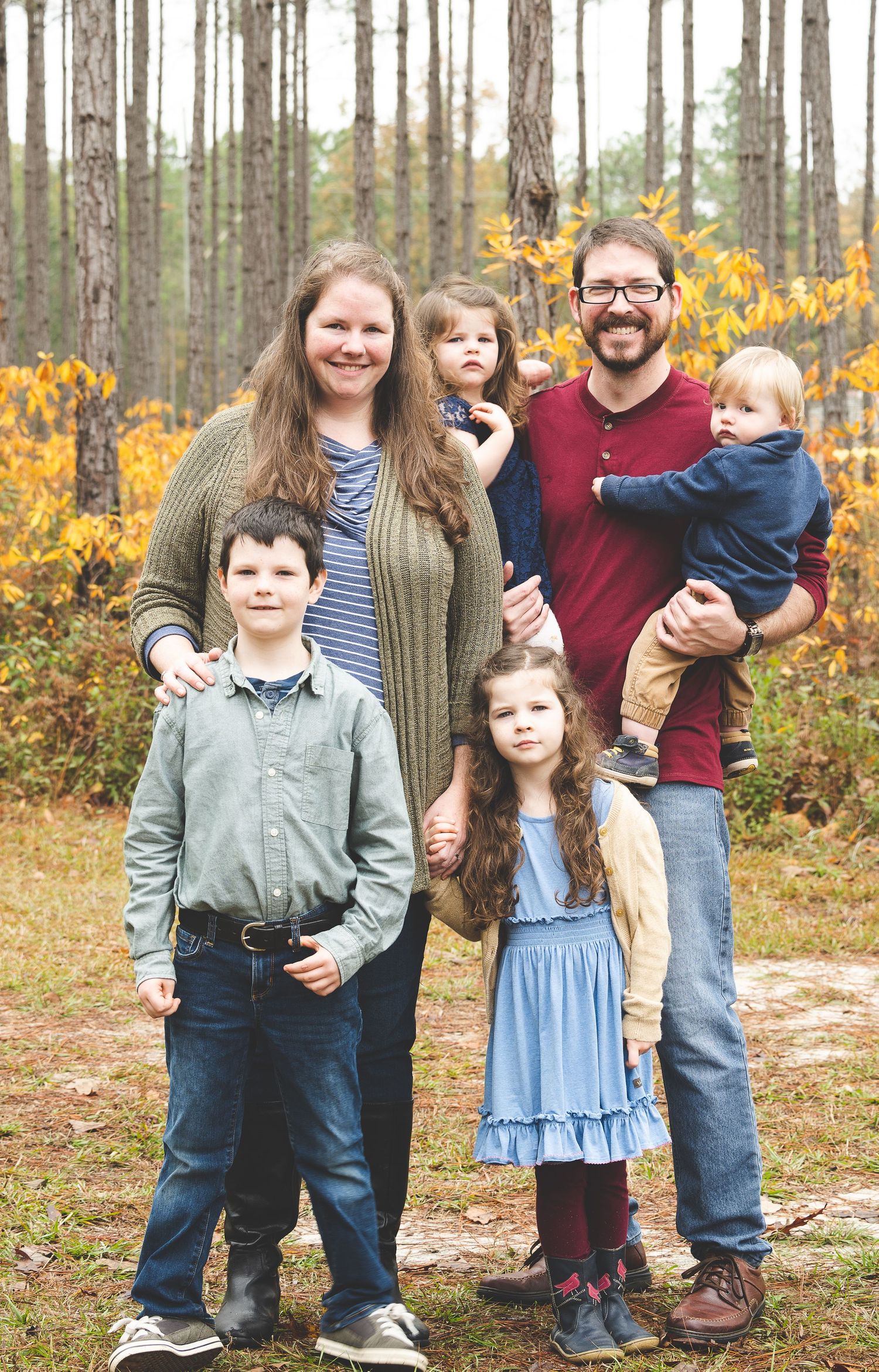 A family stands together outdoors in a wooded forest area with yellow fall foliage in the background.