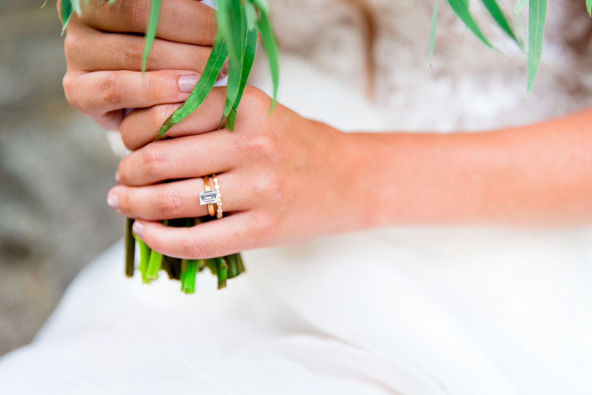 close up of hands holding a wedding bouquet with a yellow gold wedding band and emerald cut engagement ring on.