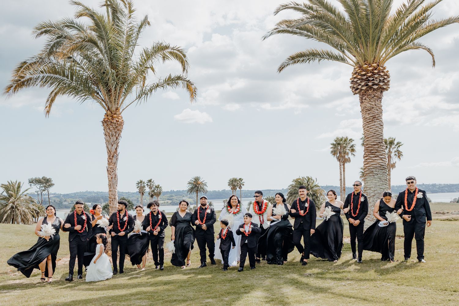 Large wedding party posing outdoors on a lawn with tropical palm trees in the background.