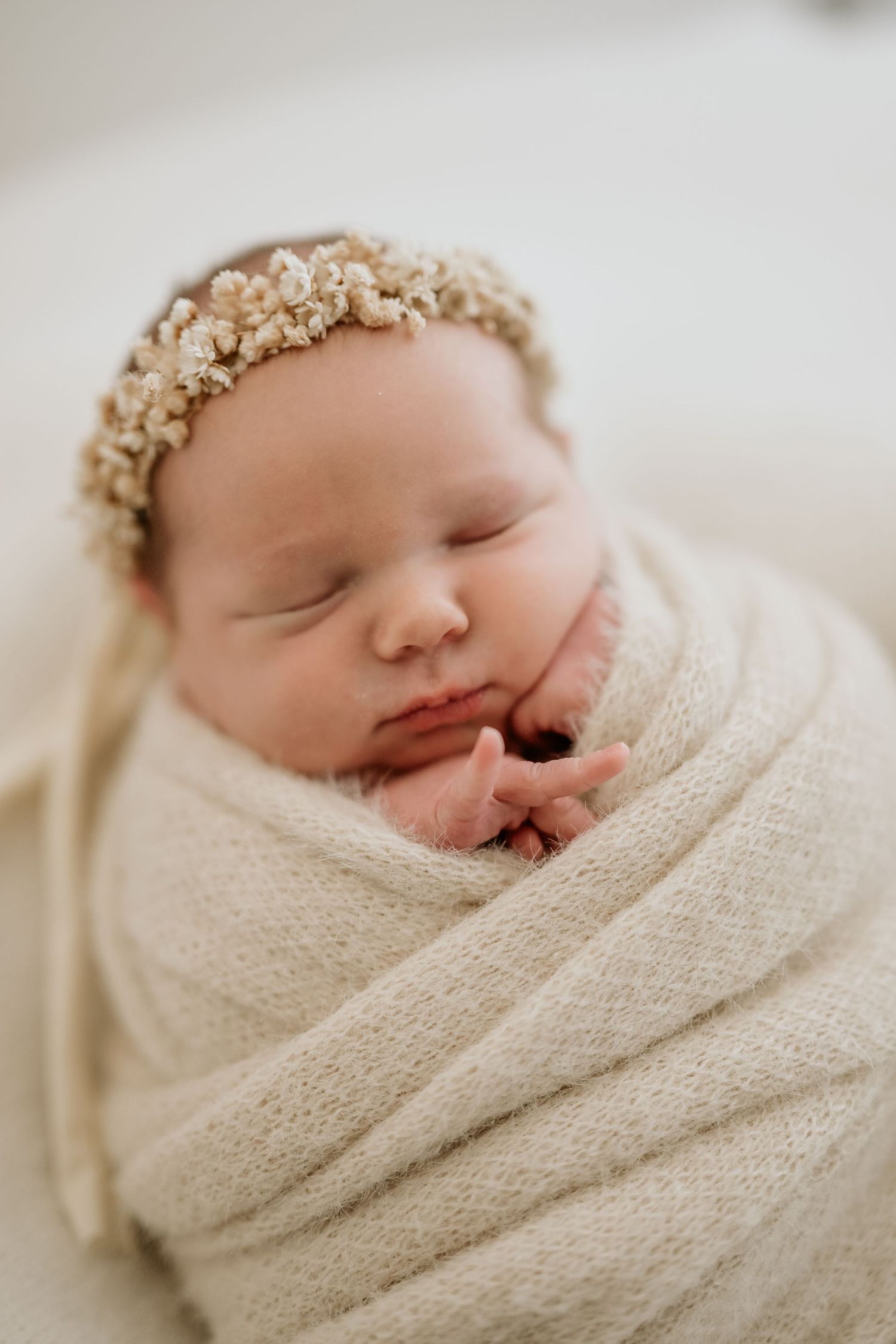 Newborn baby sleeping peacefully wrapped in cream swaddle wearing delicate floral crown.