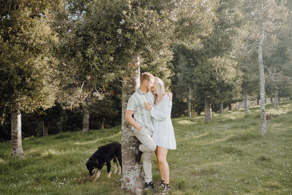 Couple poses playfully near a tree trunk while their dog explores the grassy hillside.