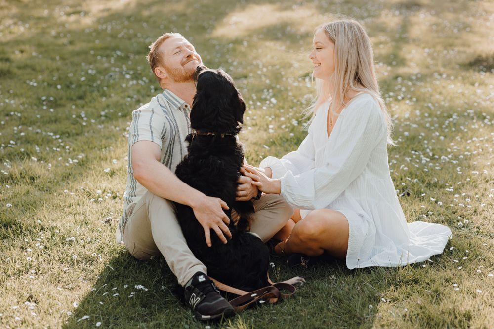 Couple sitting on grass with their black dog enjoying a sunny afternoon outdoors.