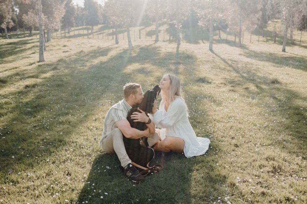 A couple sits on grass with their black dog during golden hour in a park setting.