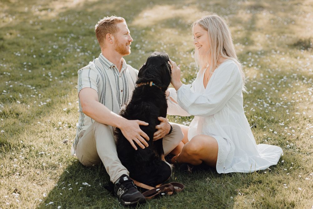 A couple sits on grass petting their black dog during a sunny afternoon outdoors.