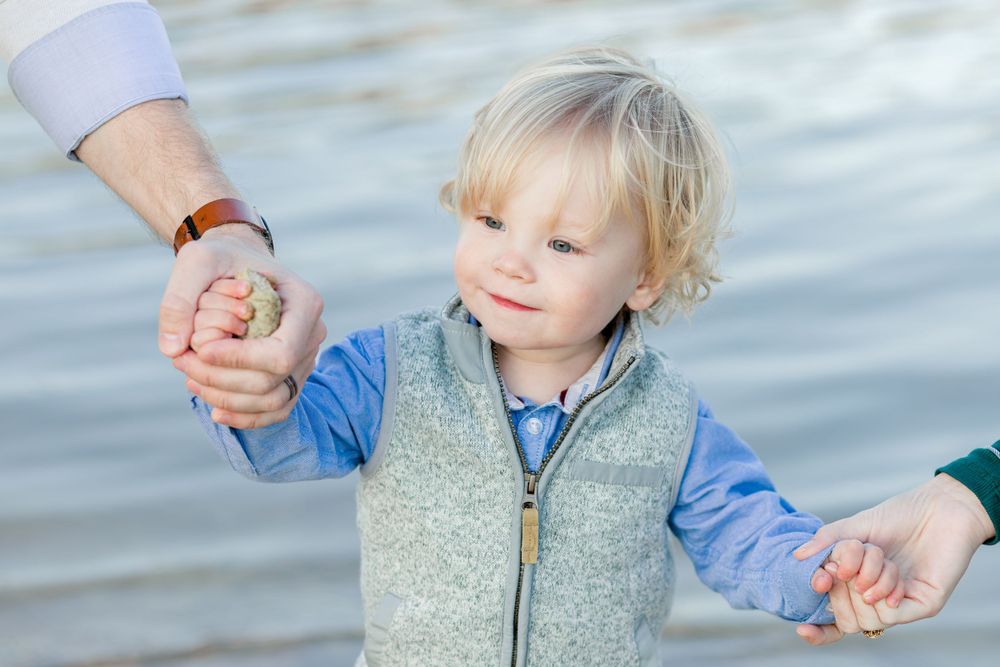 A young child holding hands with adults while wearing a blue shirt and gray vest.