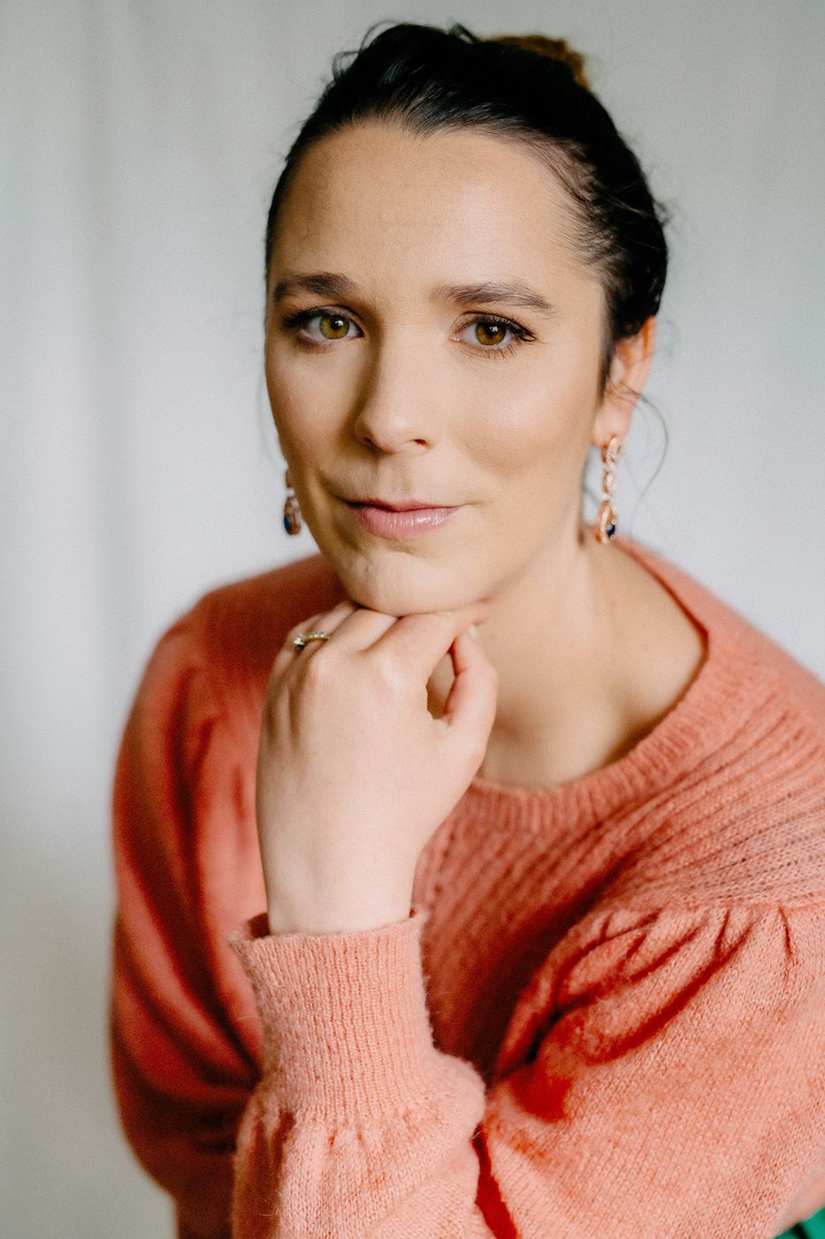 A person in an orange, textured sweater rests their chin on their hand, set against a soft, neutral background.