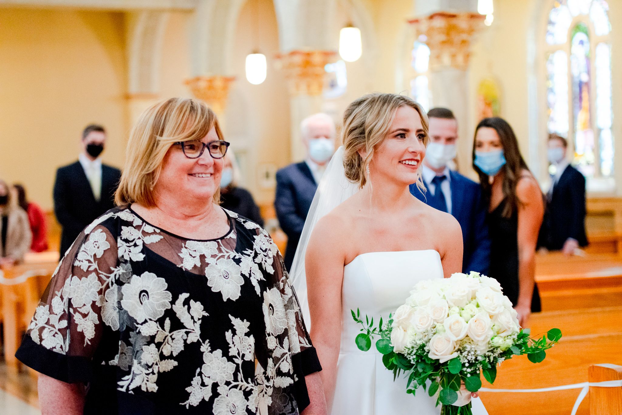 bride holding white bouquet and mother in glasses smiling as they walk down the aisle