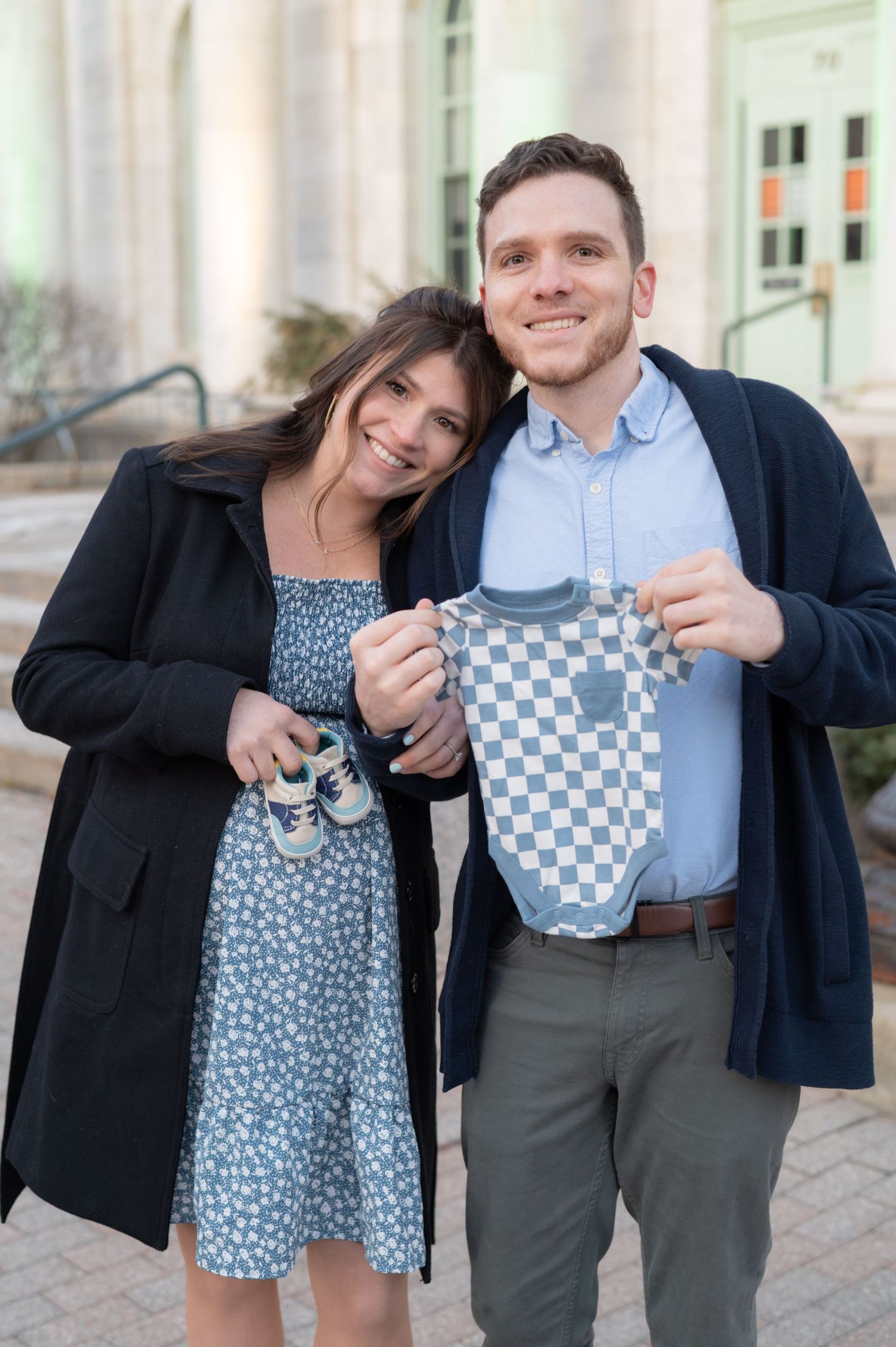 Expectant couple holding blue and white checkered baby items outside a building.
