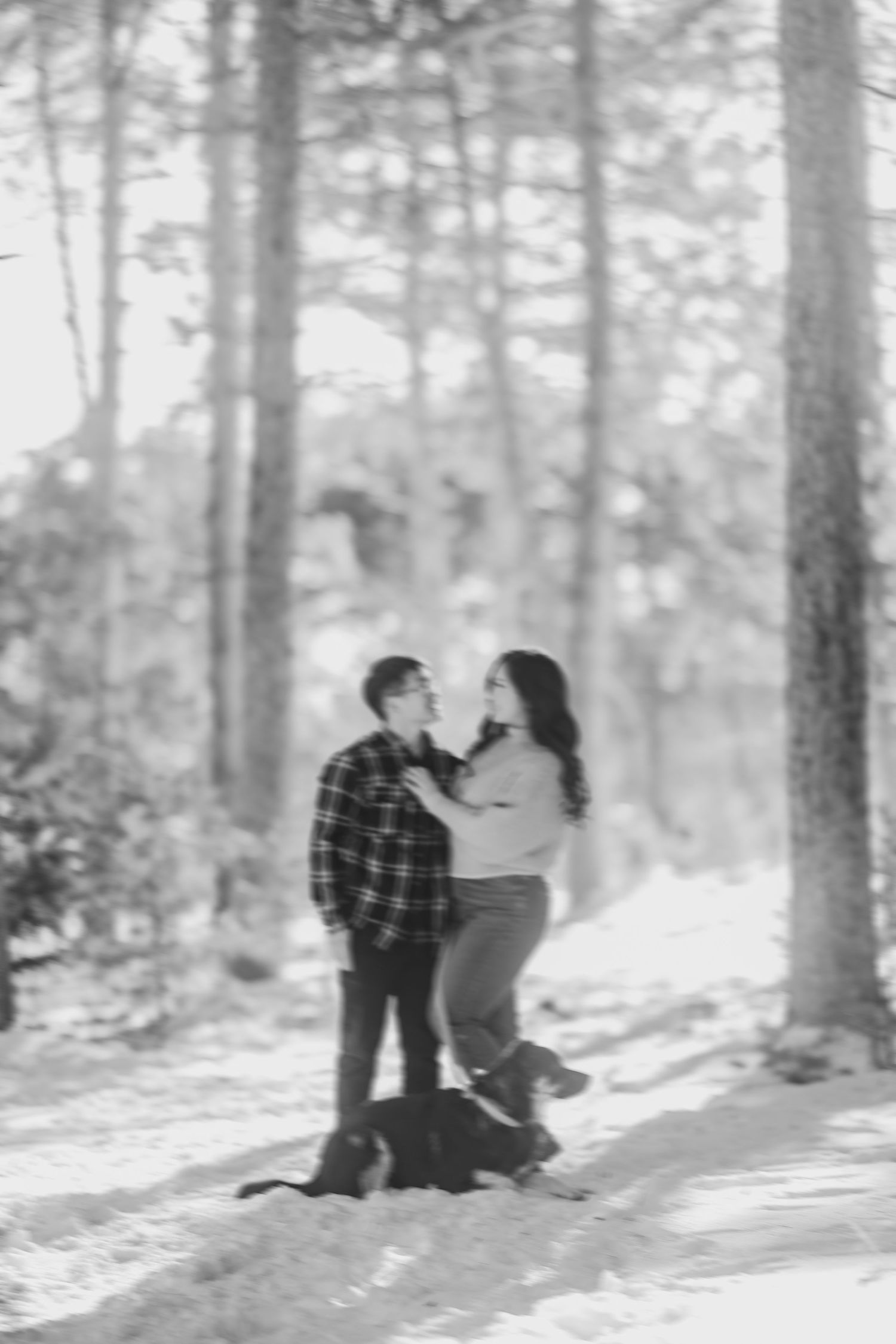 Black and white image of couple embracing on snowy forest path with dog.