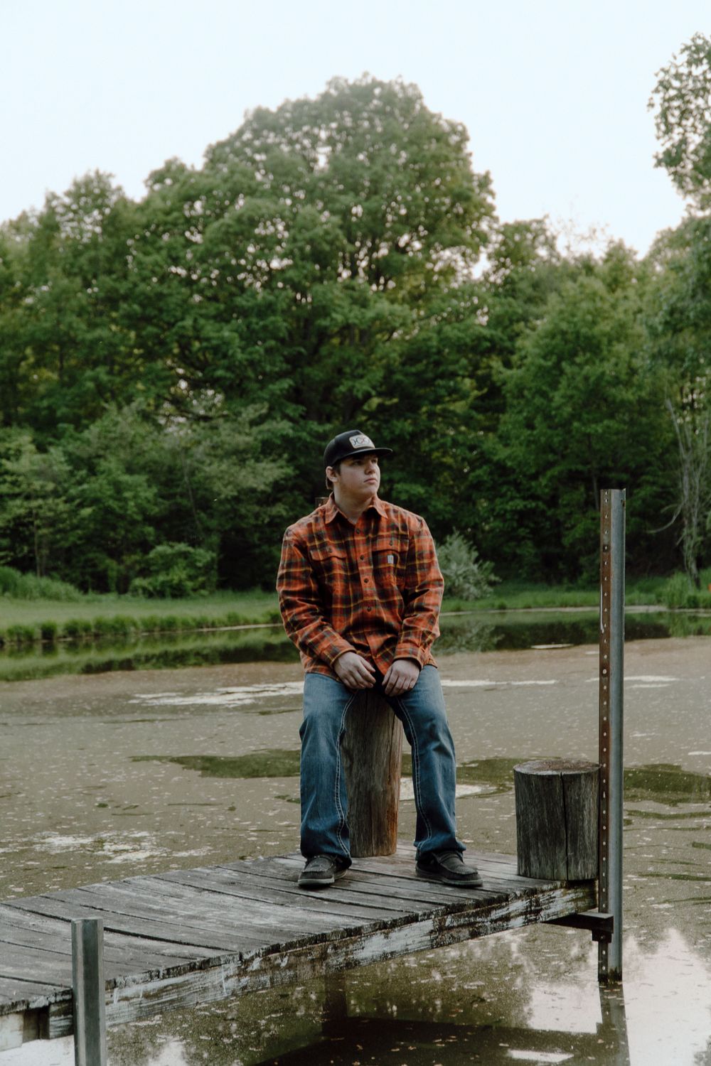 Person in plaid shirt sits on wooden dock overlooking calm water.