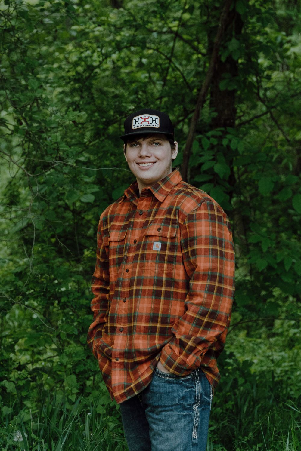 Person wearing orange plaid flannel shirt and baseball cap poses in front of green foliage.