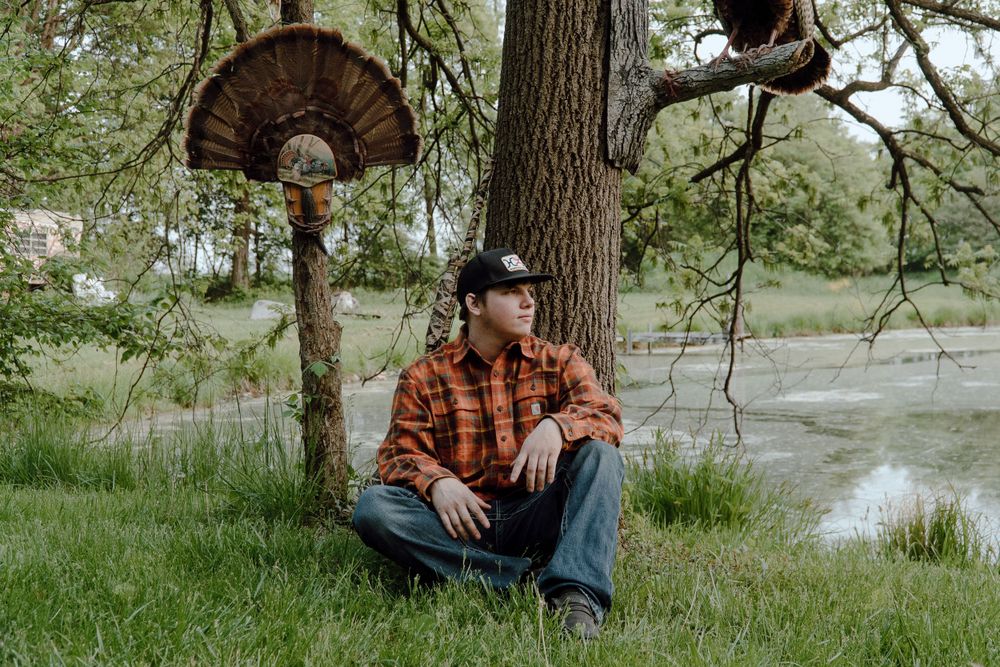 Person in orange plaid shirt sits beneath mounted turkey fan display in outdoor nature setting.