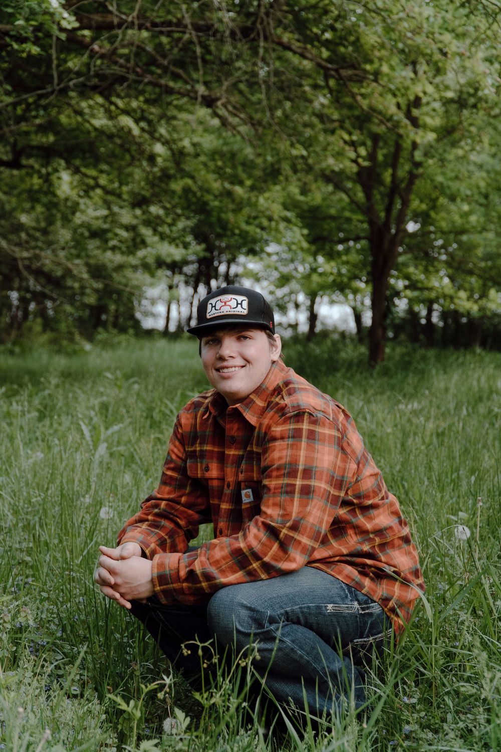 Person in plaid shirt and baseball cap crouches in green spring meadow.