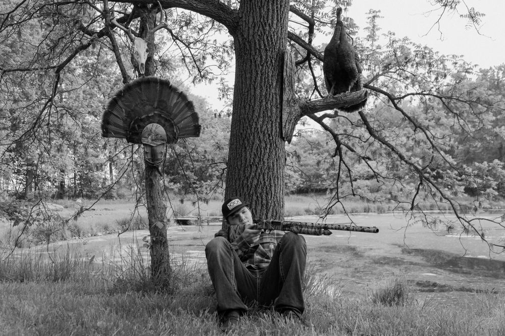 Black and white photo of someone resting against a tree trunk with displayed turkey fan mount.