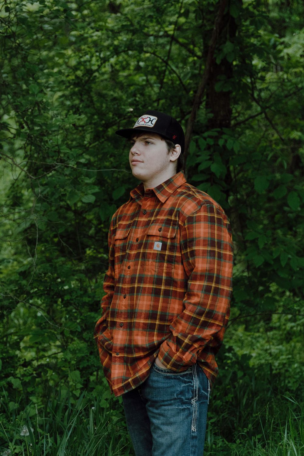 Person in plaid jacket and baseball cap stands against lush green forest backdrop.