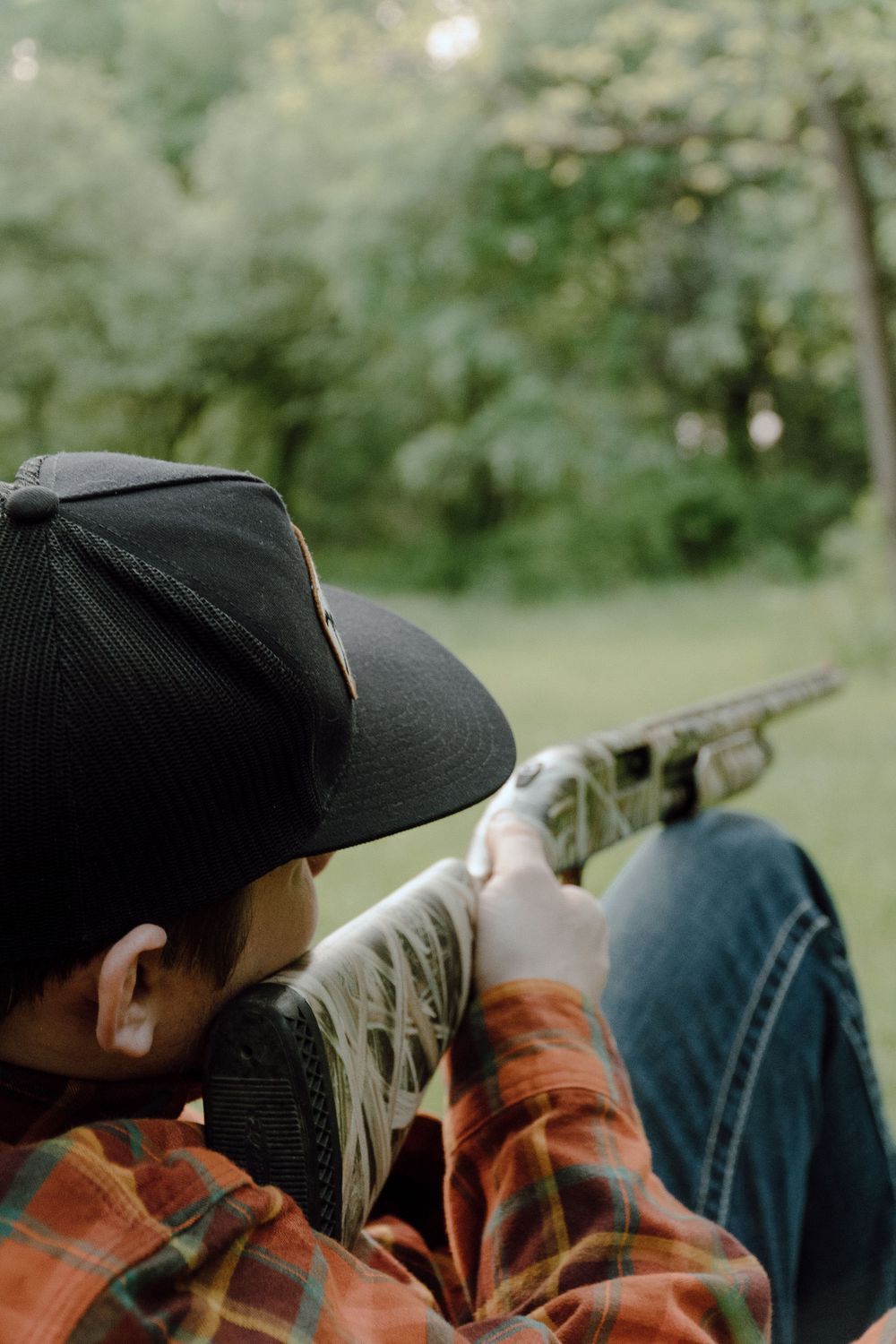 Person wearing baseball cap and plaid shirt aims shotgun in outdoor setting.