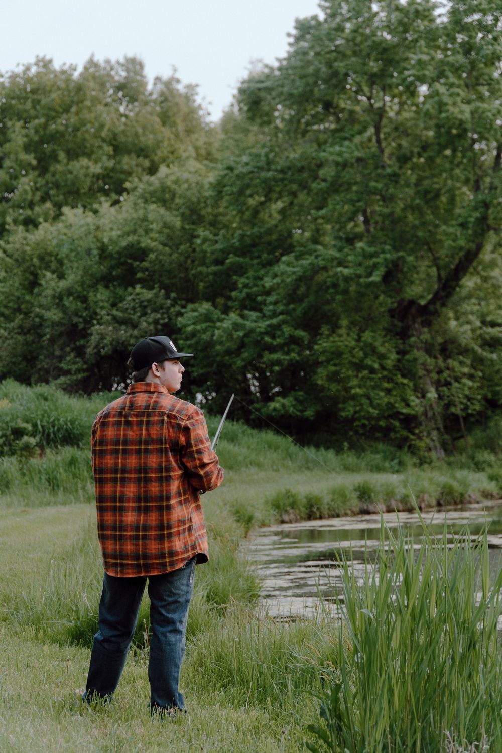 Person in orange plaid shirt stands fishing by peaceful pond with cattails.