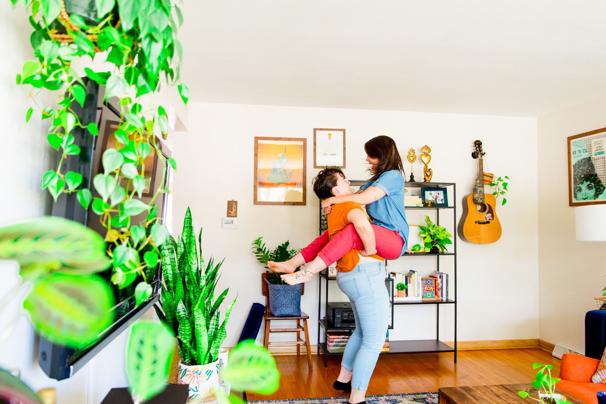 woman in orange shirt picking up woman in blue shirt and pink pants in front of wall with plants and guitar hanging
