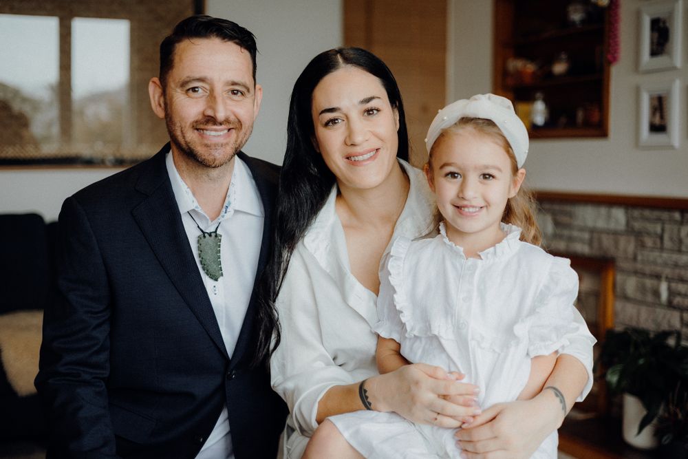 Family portrait showing couple in formal attire with young child in white dress sitting together in a living room.