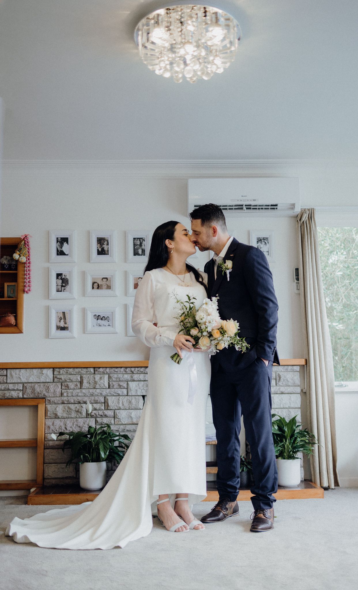 A wedding couple embraces in an elegant room with crystal chandelier and framed photos.