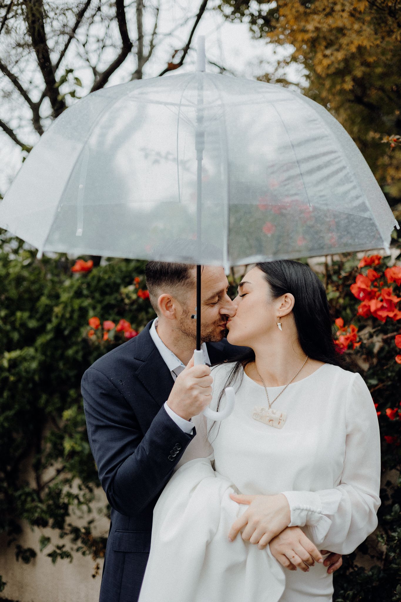 A couple shares an intimate moment under a clear umbrella with red flowers in background.