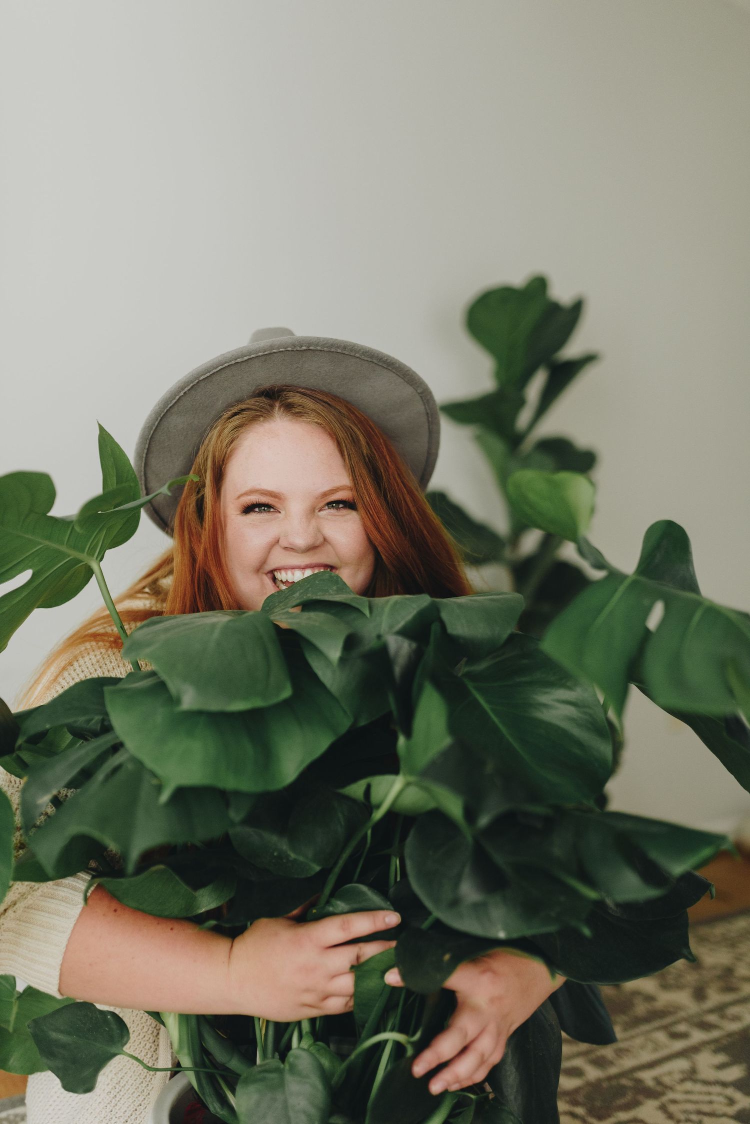 Natural light photo of woman kneeling on the ground with arms around a monstera deliciosa