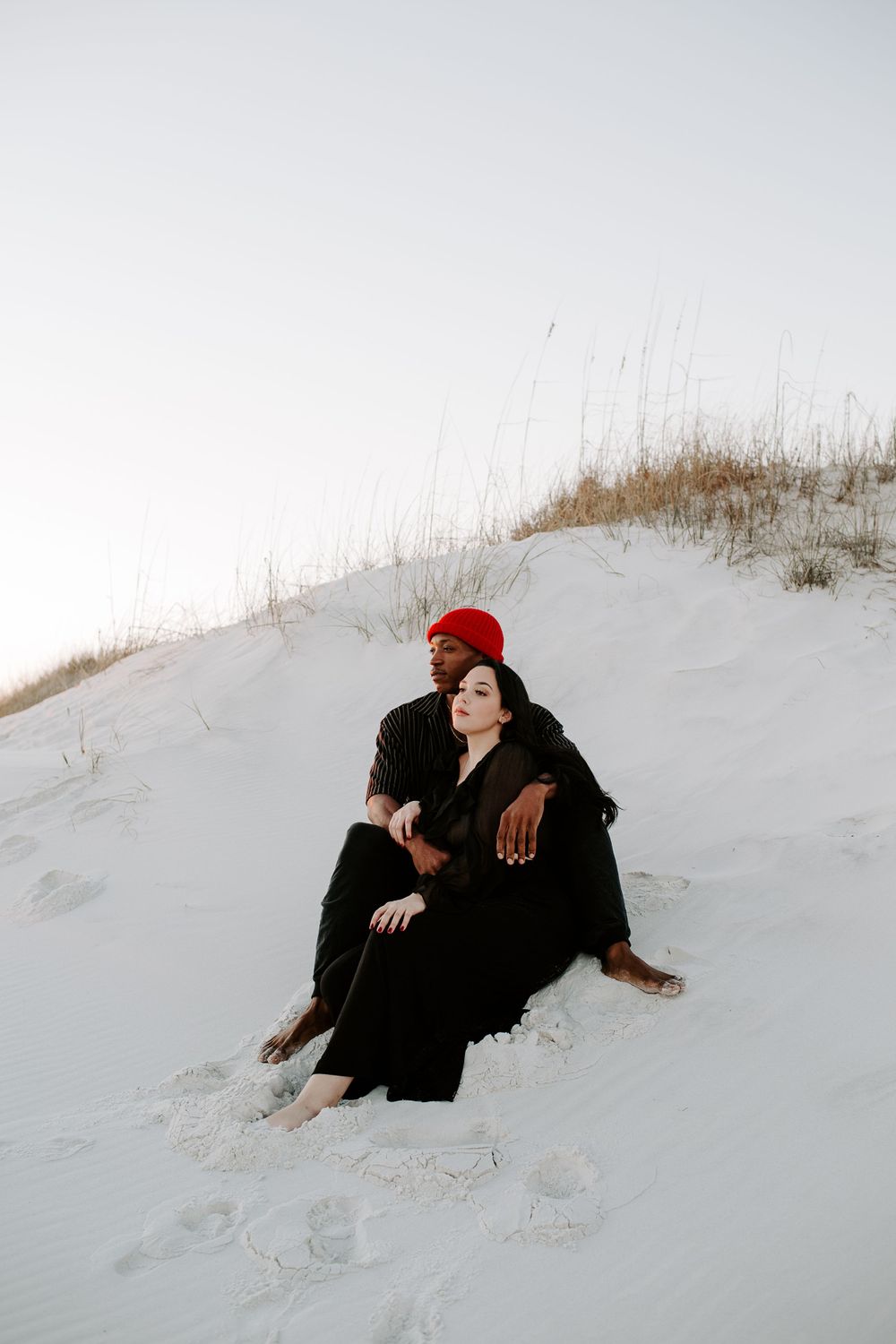 couple sitting near sand dunes looking off into the distance during a sunset photoshoot
