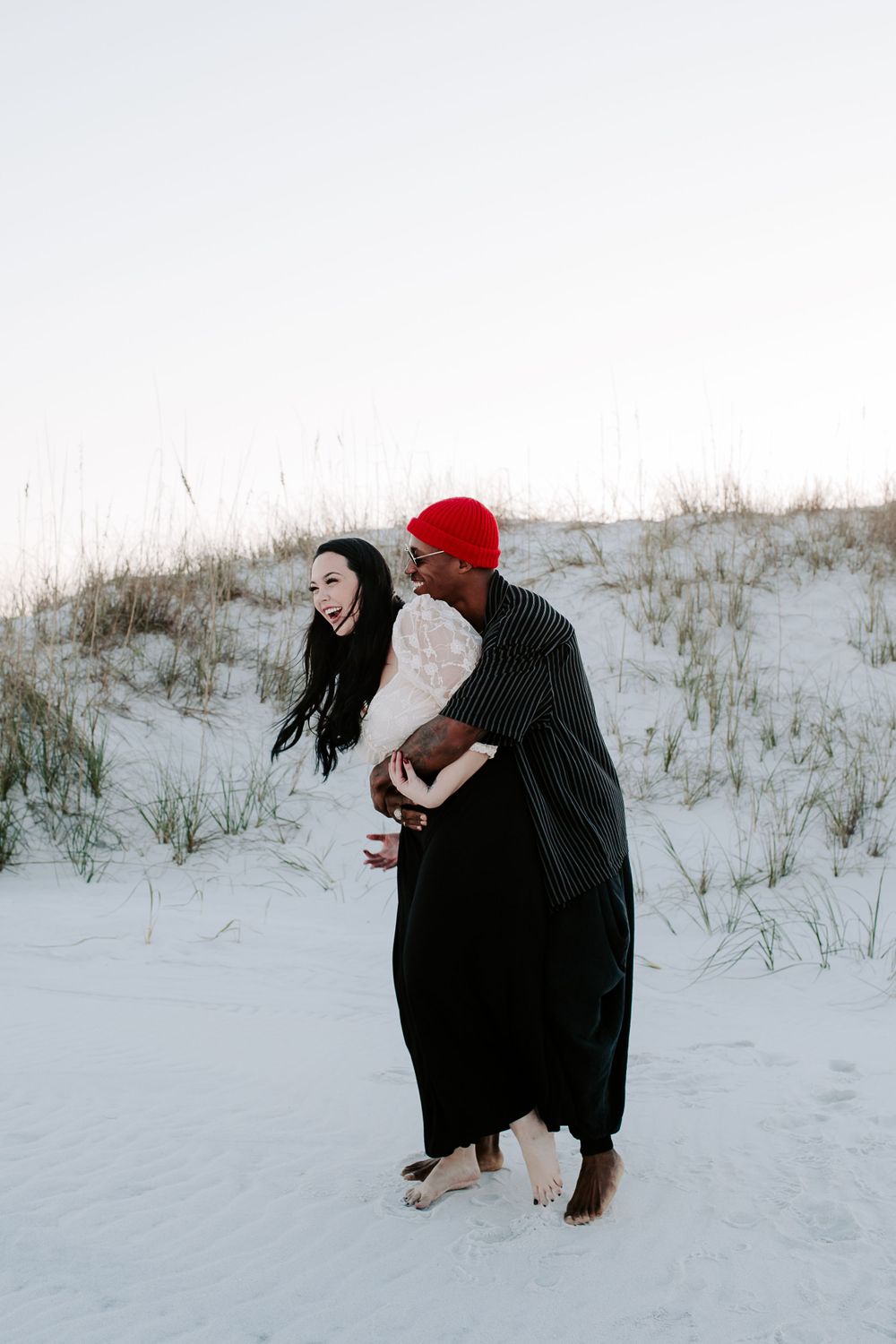 man embracing a woman from behind in front of sand dunes during sunset