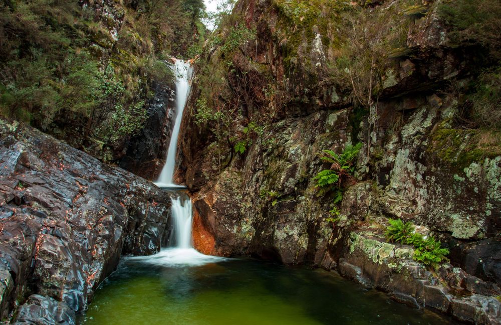 A scenic waterfall cascades down rocky cliffs into a green pool surrounded by moss-covered rocks and vegetation.