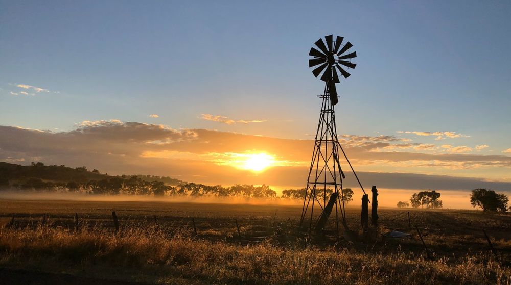 Golden sunset over rural farmland with a silhouetted windmill against the orange sky.