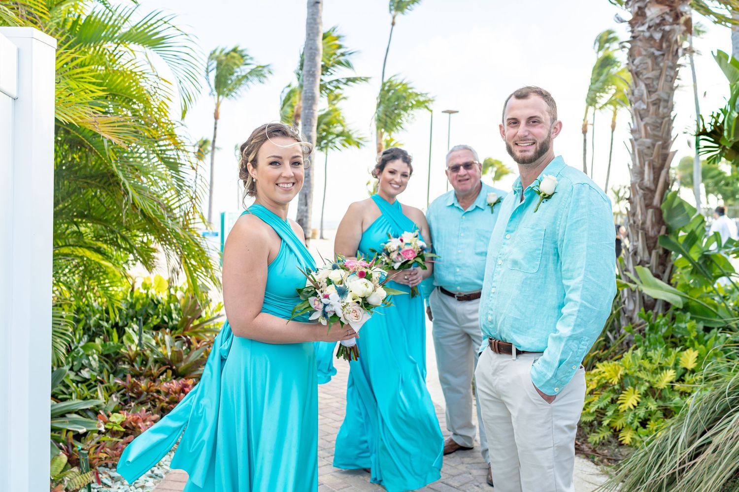 Wedding party members in turquoise and white attire pose outdoors among tropical palm trees.