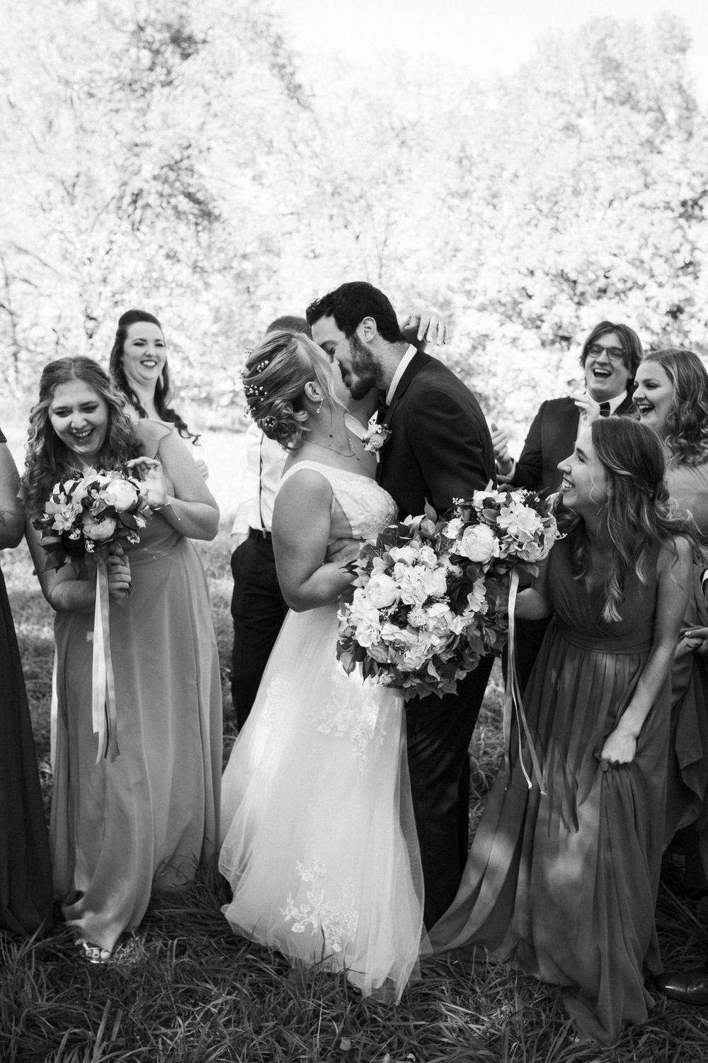 A bride and groom kissing while their wedding party surrounds them and smiles at them.