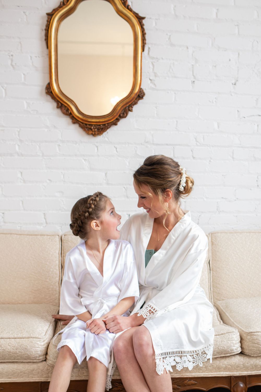 Two people in white robes share an intimate moment while sitting on a cream couch beneath an ornate gold mirror.