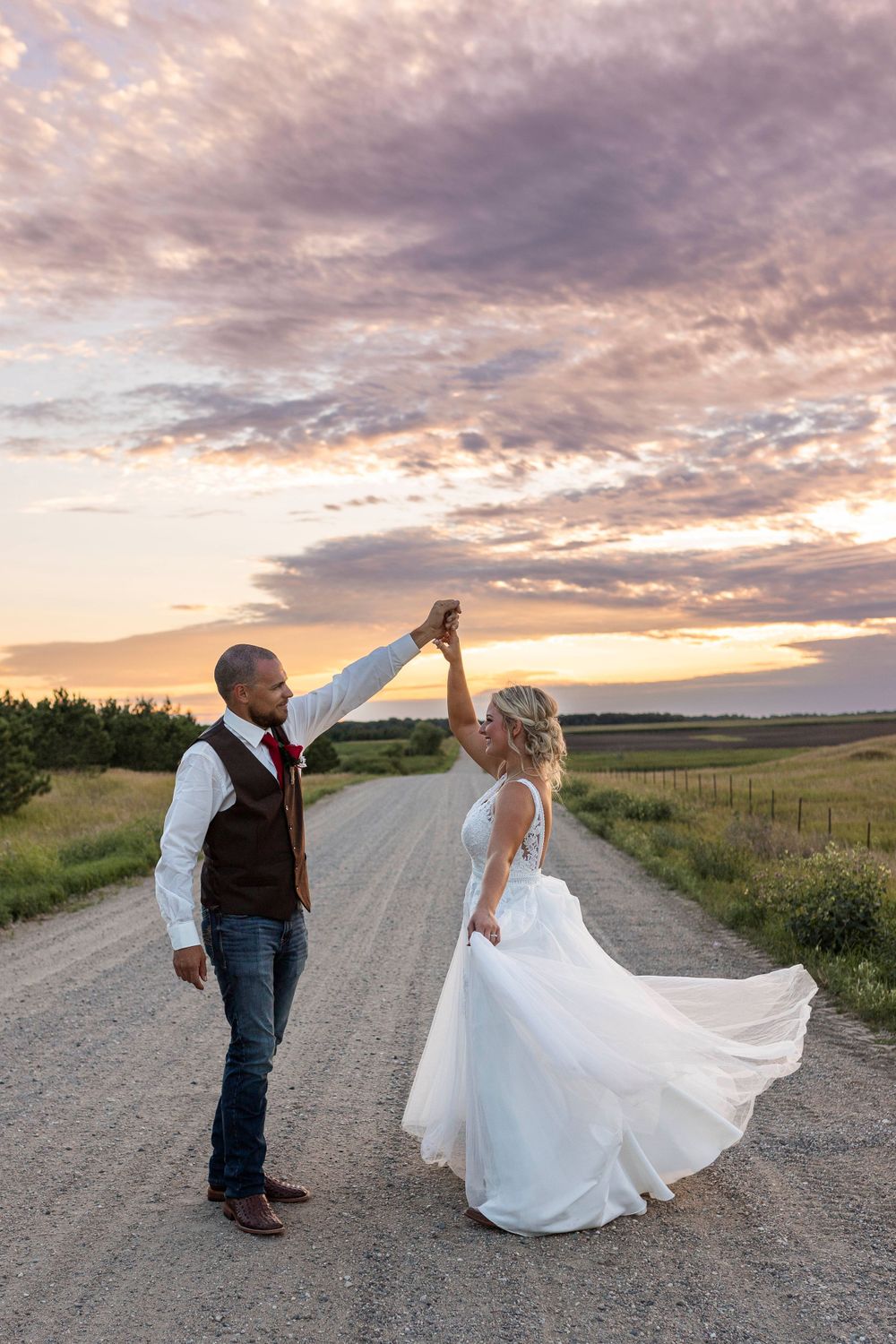 A groom twirling his bride on a dirt road with the sun setting in Central, MN.
