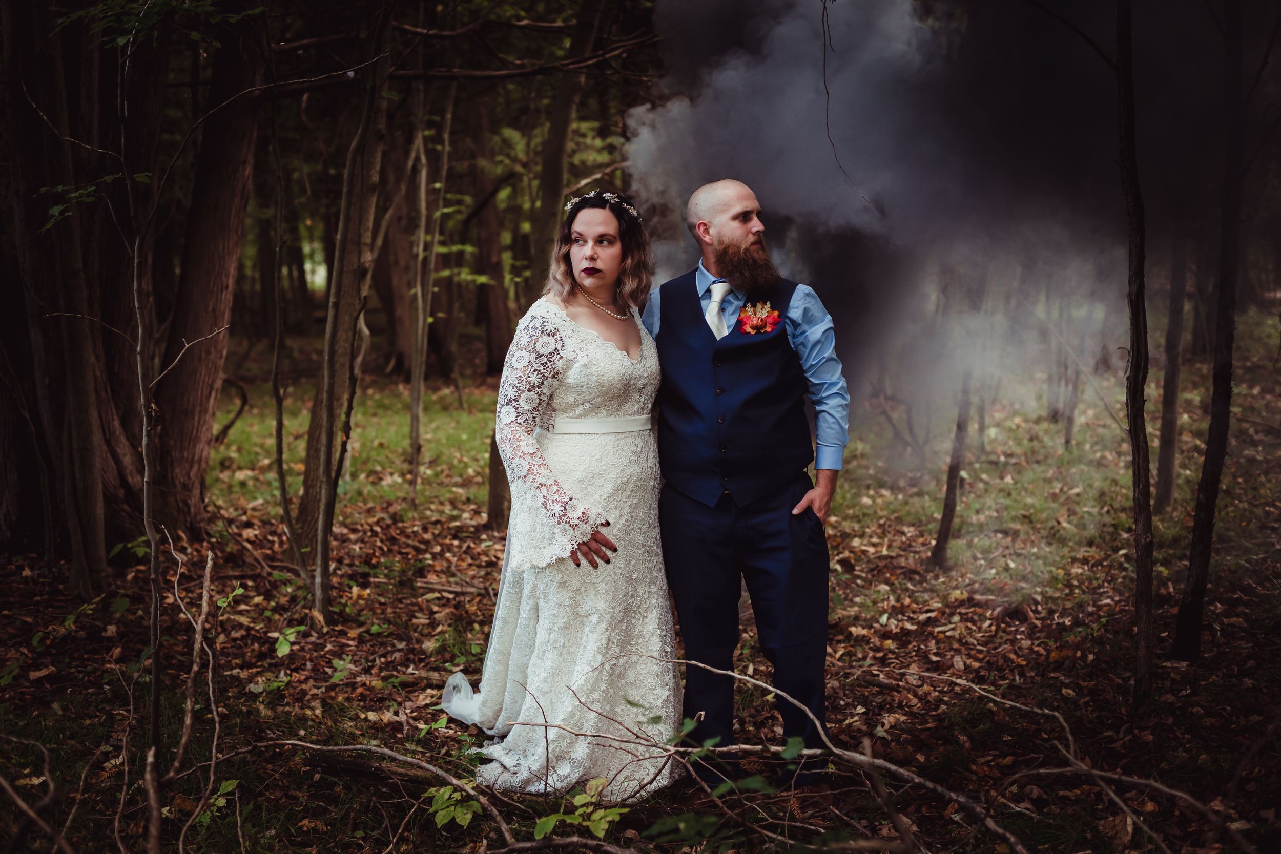 Woman and man in wedding attire standing in the woods with black smoke behind them.