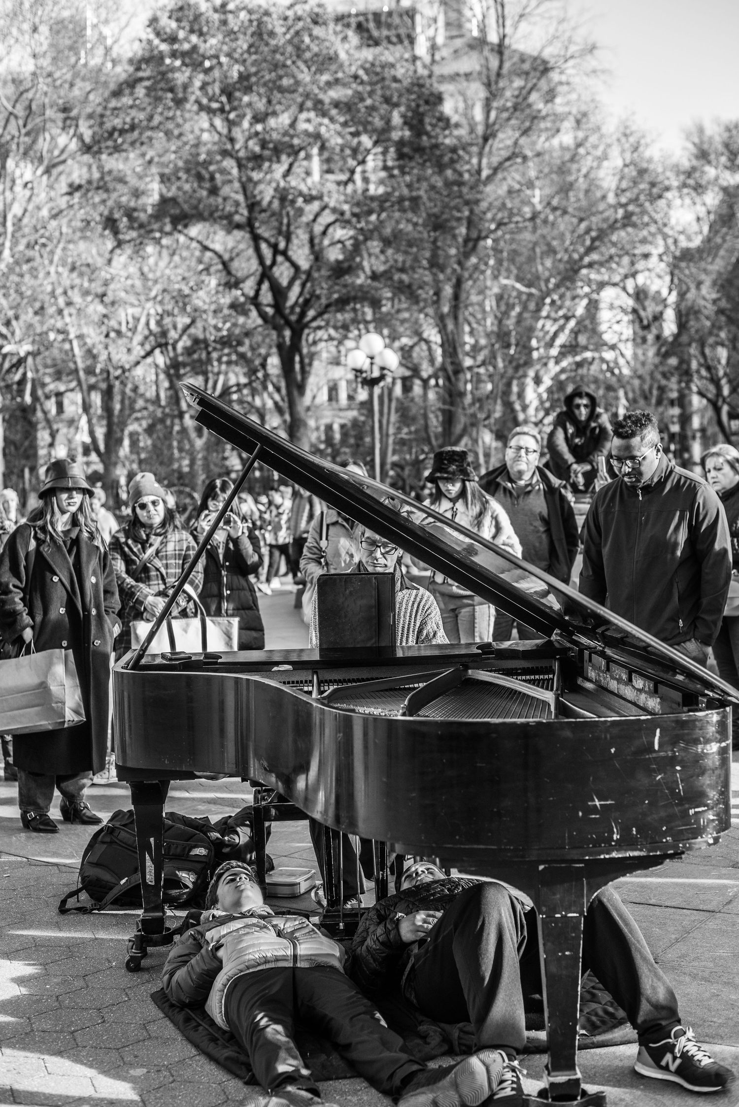 Two men lying under piano while pianist performs in Washington Square Park, NYC