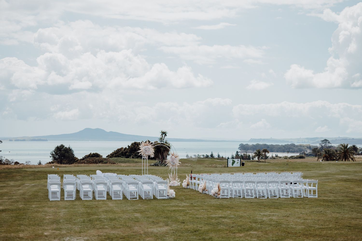 White wedding chairs set up in rows on a scenic oceanfront lawn for an outdoor ceremony.