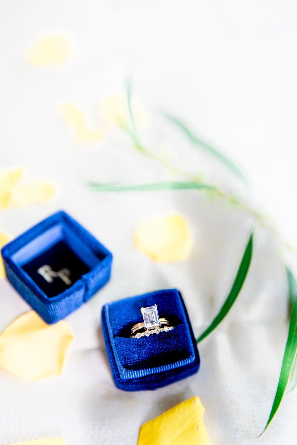 emerald cute diamond engagement ring and gold wedding ring in blue ring box next to greenery and yellow rose petals