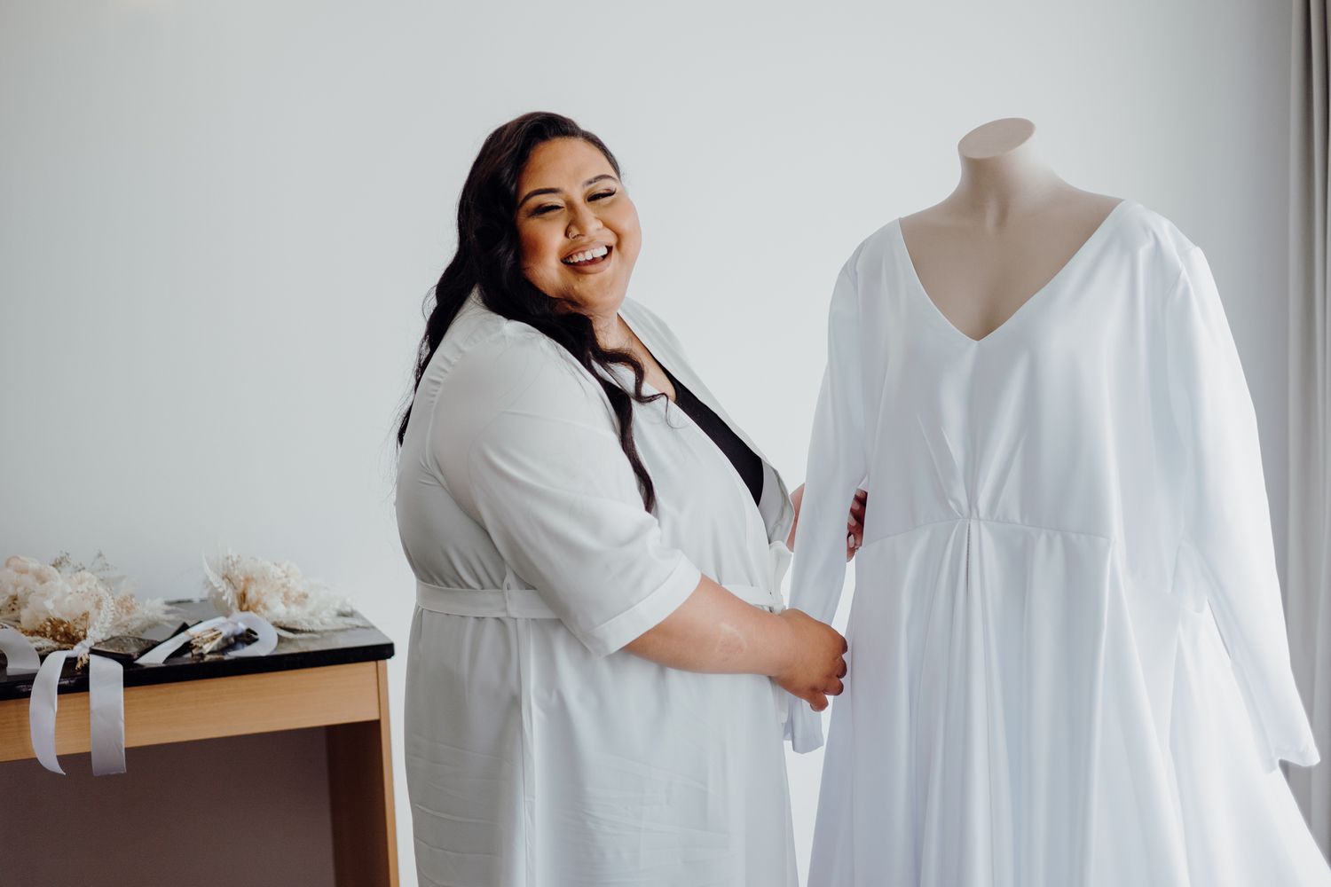 Person in white robe smiling while standing next to wedding dress on mannequin.