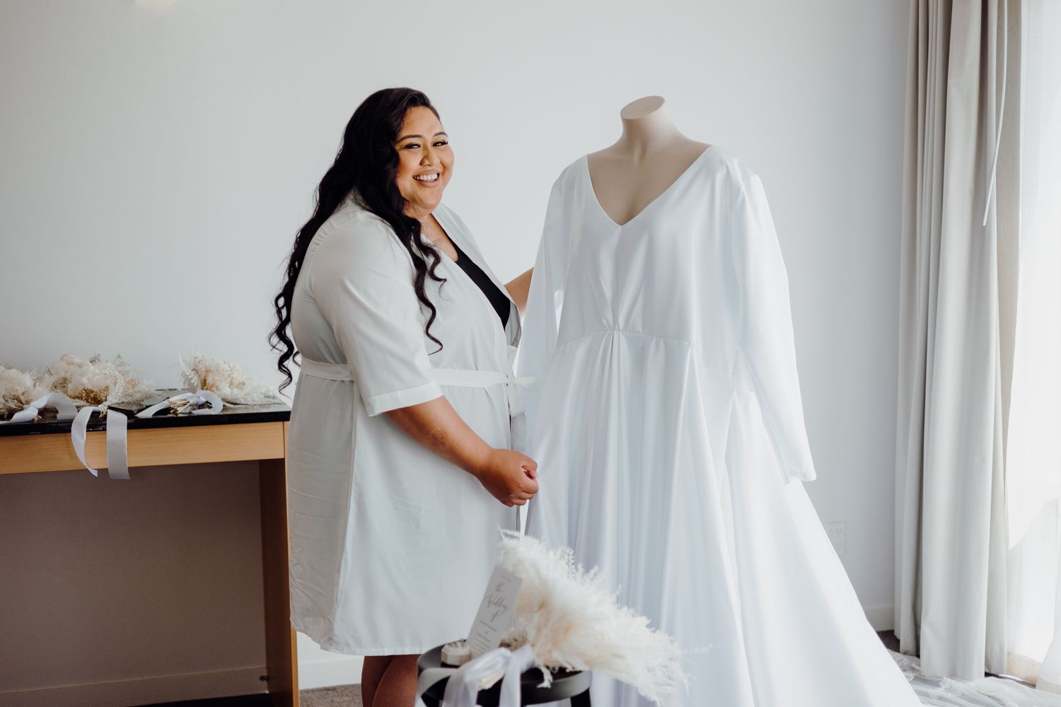 Person in white robe admiring wedding dress displayed on mannequin.