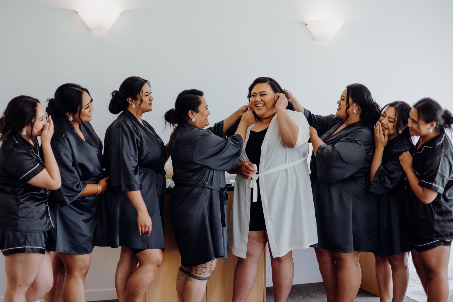 Wedding party in matching black dresses surrounding person in white dress during preparation.
