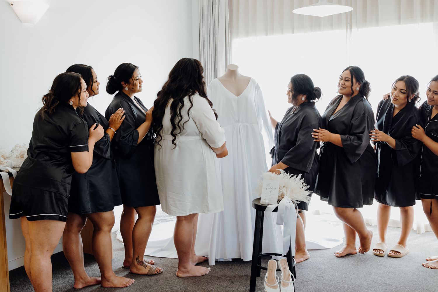 Group gathered around hanging white wedding dress in preparation room.