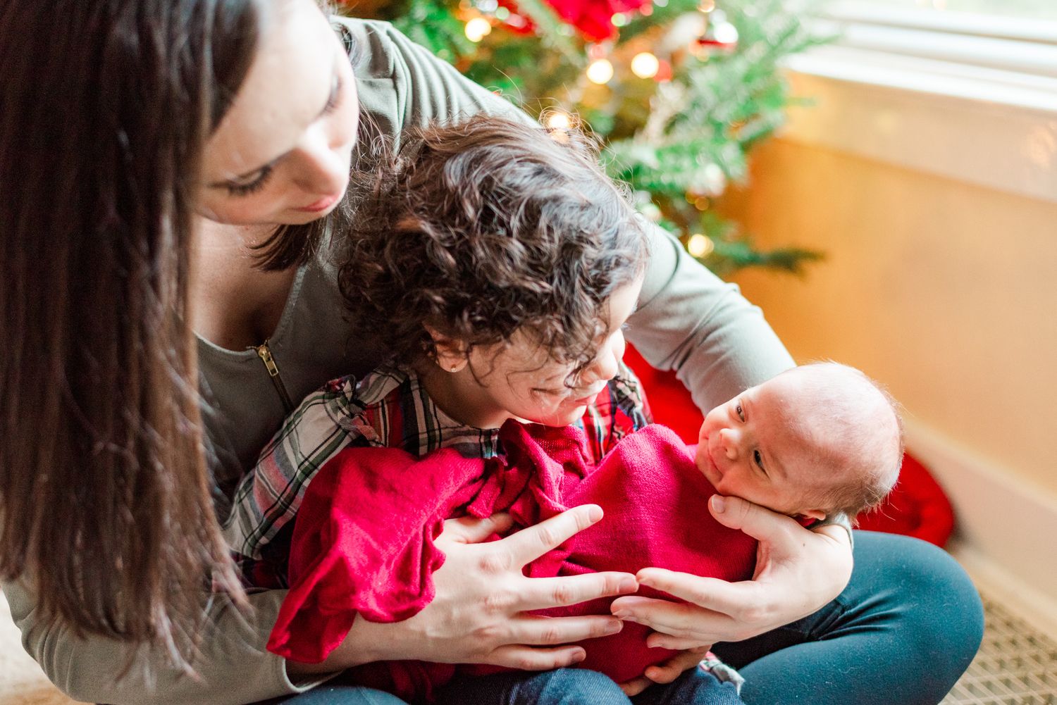 Lifestyle mother and big sister holding newborn baby in pink wrap in front of Christmas tree in Pittsburgh, Pennsylvania