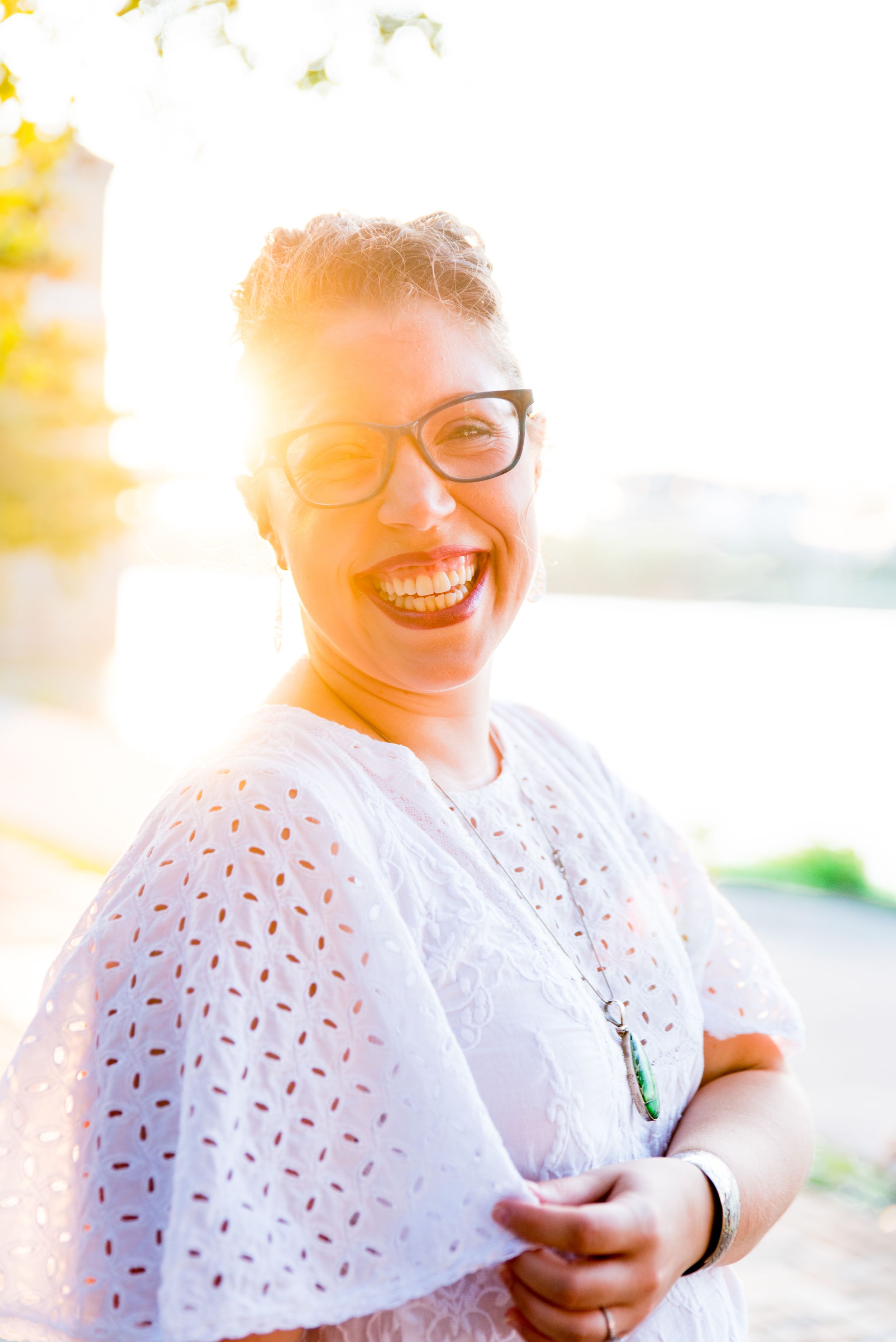 woman in white shirt, red lipstick, and glasses laughs and smiles in front of Roebling Bridge at sunset