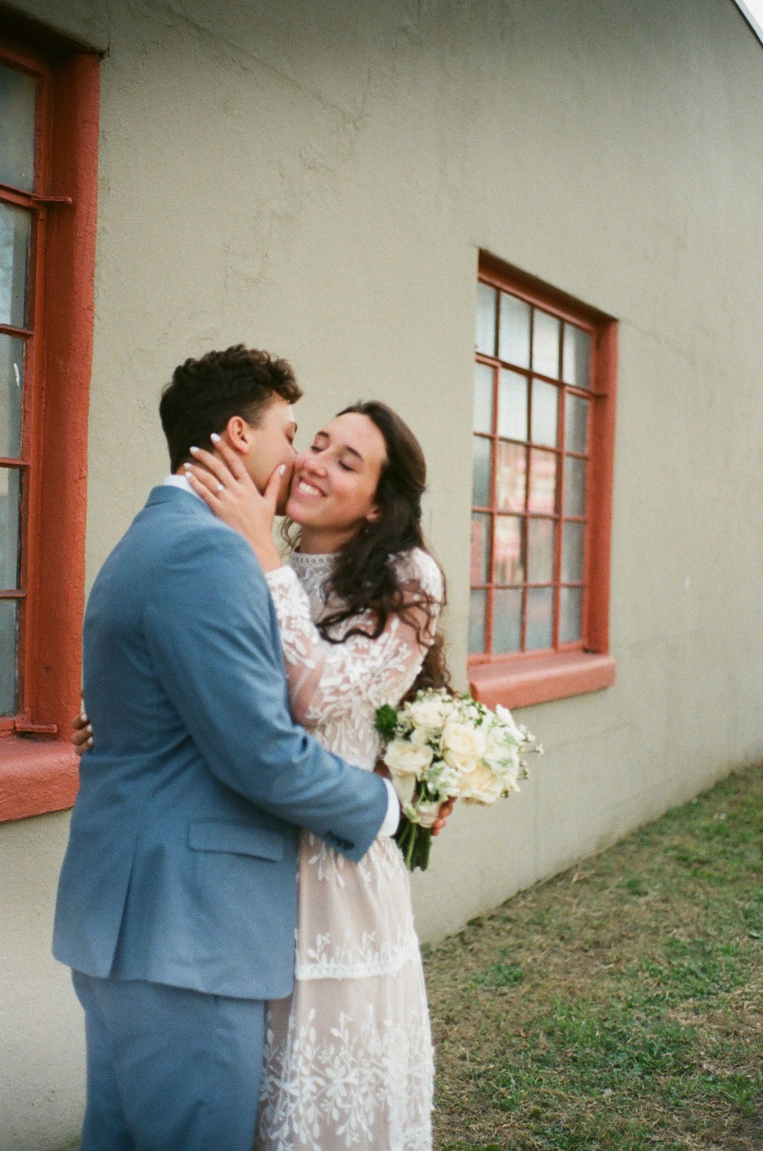 A couple in wedding attire shares a romantic moment against a white wall with red-trimmed windows.