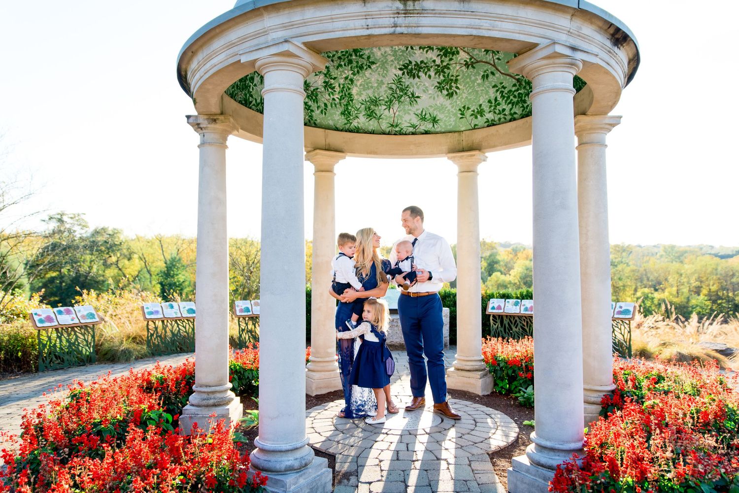mom and dad smiling at each other and holding kids and one kid is hugging mom's knee under a gazebo at sunset