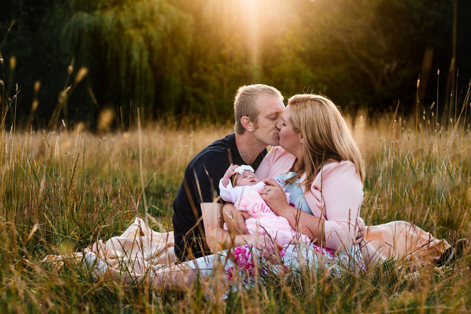In a field in Havant at sunset new parents sit with their newborn daughter, kissing in the long grass.